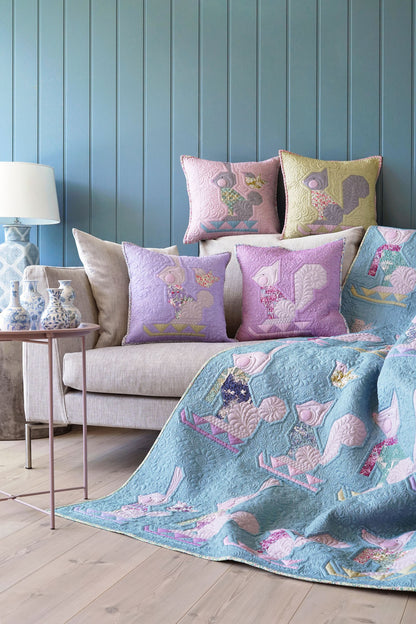 A cozy living room scene featuring a light blue quilt with pink and purple quilted rabbits draped over a beige sofa, surrounded by matching decorative pillows against a blue paneled wall.
Photo by Tildafabrics.com [additional_image_link]
