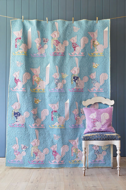 A light blue quilted blanket featuring a repeating pattern of pink and lavender squirrels, hanging on a clothesline in a room. A white wooden chair with a matching lavender pillow sits in front of the quilt against a dark blue paneled wall.
Photo by Tildafabrics.com [additional_image_link]