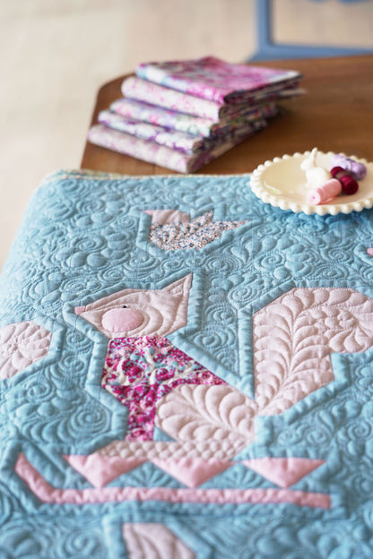 A close-up of a quilt featuring a stylized fox design in light blue and pink, with floral patterned accents. A stack of folded matching fabrics and a small dish with beads sit on a wooden table in the background.
Photo by Tildafabrics.com [additional_image_link]