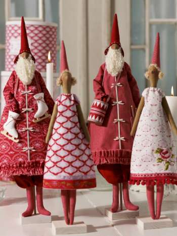 Four Christmas gnome figurines in red and white holiday clothing, standing on small white pedestals in front of a softly blurred indoor background with candles.
Photo by Tildafabrics.com [additional_image_link]
