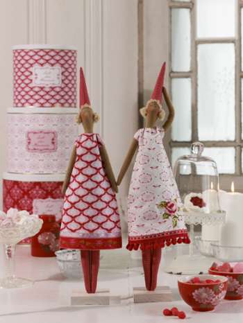 Two decorative gnome figurines in red and white patterned dresses holding hands, displayed on a tabletop with red patterned boxes, candles, and glassware in a softly lit indoor setting.
Photo by Tildafabrics.com [additional_image_link]
