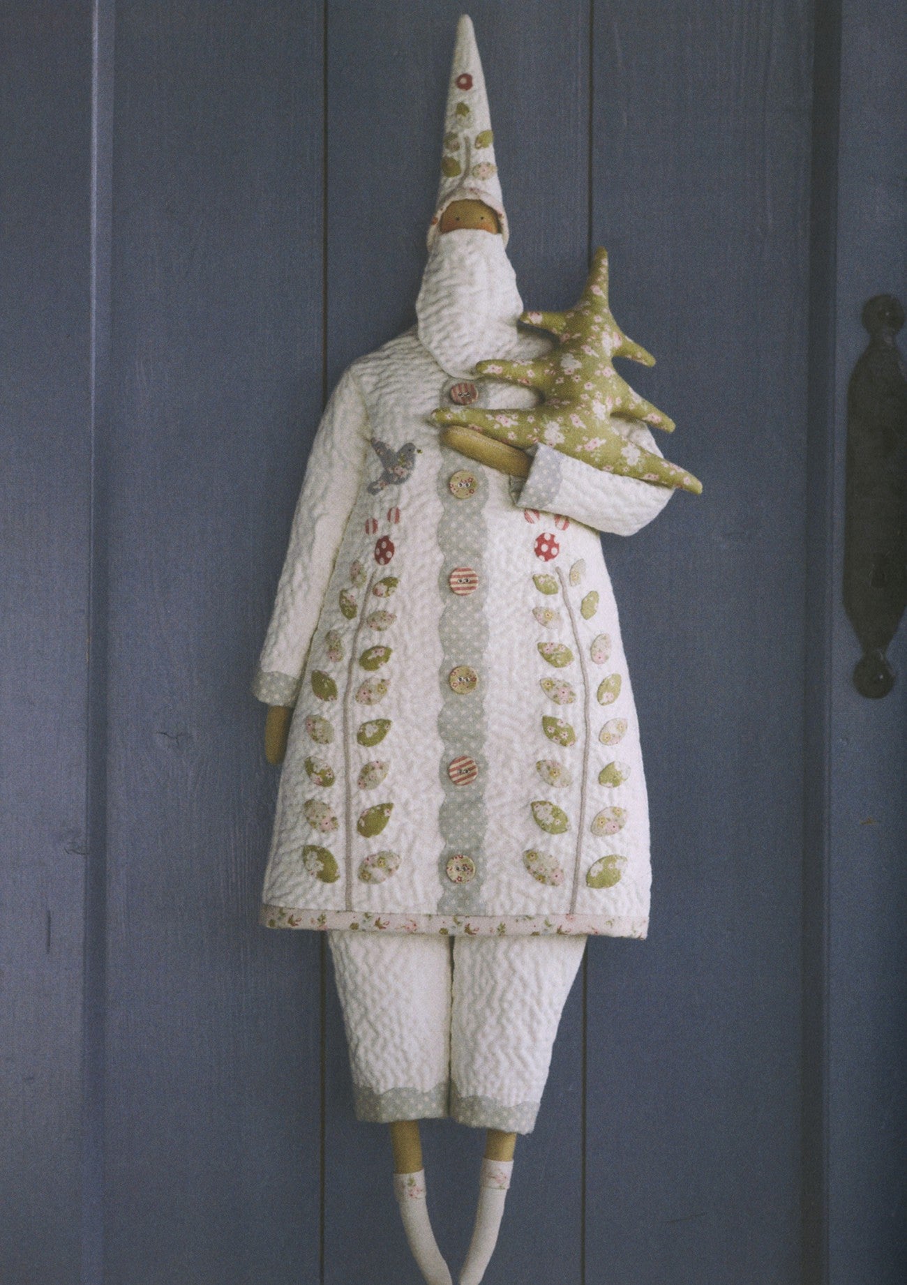 A white knitted Christmas gnome doll with a tall pointed hat, holding a small gold Christmas tree, displayed against a dark blue-gray paneled door.
Photo by Tildafabrics.com [additional_image_link]