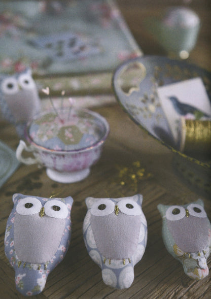 Three small owl-shaped decorative ornaments or figurines in soft pastel fabric, arranged on a wooden surface with a blurred vintage-style teacup and floral background.
Photo by Tildafabrics.com [additional_image_link]