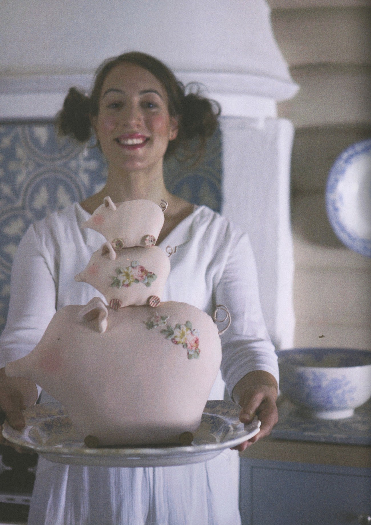A smiling woman in a white dress holds a decorative ceramic pig-shaped serving piece on a silver platter in a softly lit indoor setting.
Photo by Tildafabrics.com [additional_image_link]