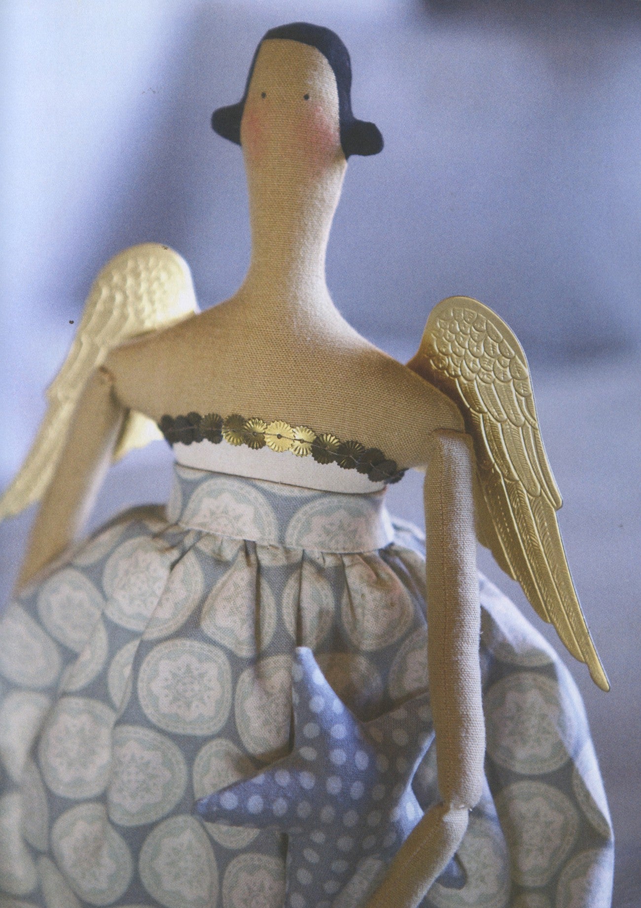 A handmade angel doll with a long neck, black hat, gold wings, and a patterned dress, shown against a soft blue-gray background.
Photo by Tildafabrics.com [additional_image_link]