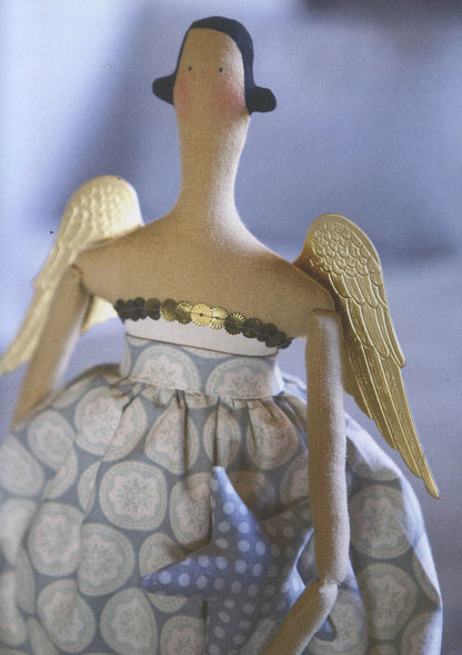 A handmade angel doll with a long neck, black hat, gold wings, and a patterned dress, shown against a soft blue-gray background.
Photo by Tildafabrics.com [additional_image_link]