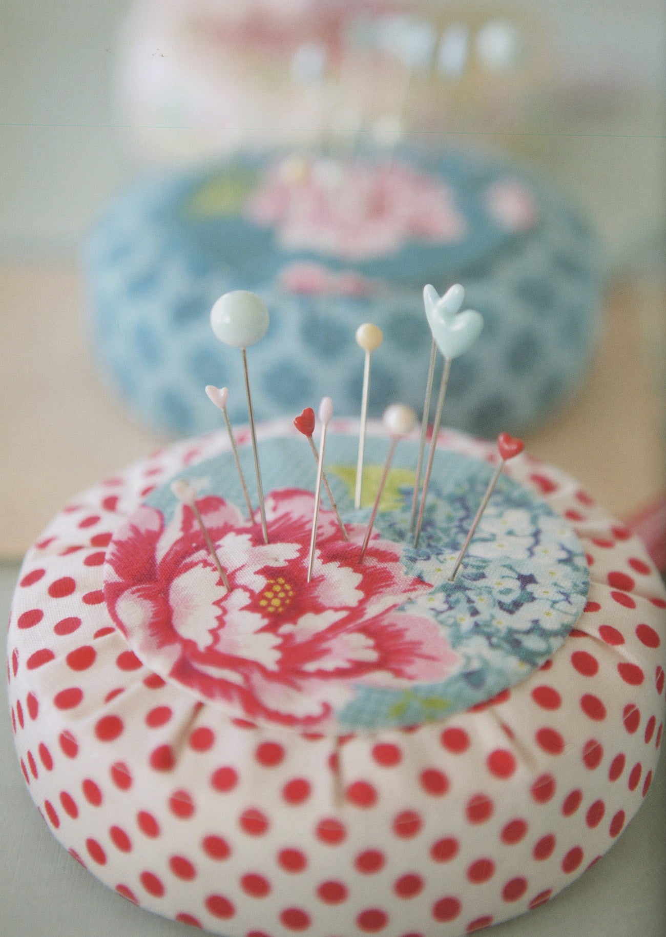 A close-up of a round pin cushion with a red polka dot pattern on the outside and a floral fabric insert on top, holding several sewing pins. A second pin cushion is blurred in the background.
Photo by Tildafabrics.com [additional_image_link]