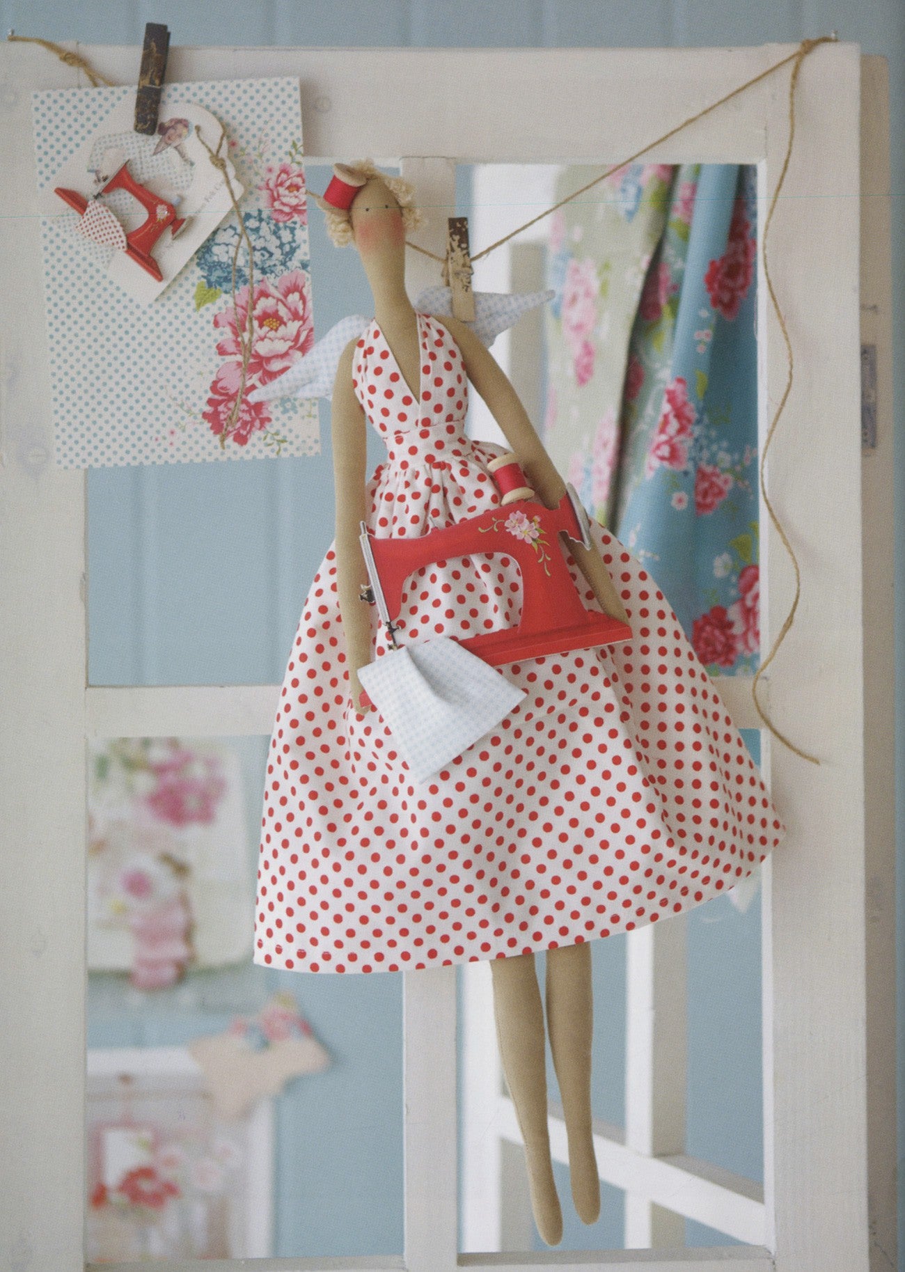 A decorative doll with a red and white polka dot dress and a red sewing machine accessory, hanging in front of a white window frame with floral fabric and paper clips.
Photo by Tildafabrics.com [additional_image_link]