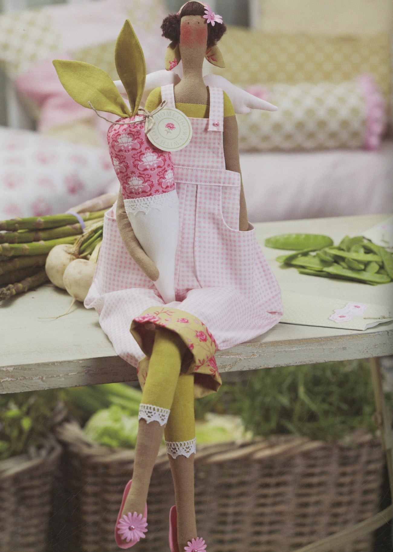 A whimsical handmade doll dressed in a pink gingham dress and yellow tights, holding a pink patterned carrot, posed on a rustic table with vegetables in a soft, blurred garden setting.
Photo by Tildafabrics.com [additional_image_link]