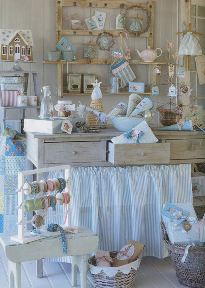 A styled vintage-inspired craft or sewing room display featuring a light wood desk with a blue and white striped fabric skirt, surrounded by sewing supplies, yarn spools, baskets, and small decorative items.
Photo by Tildafabrics.com [additional_image_link]