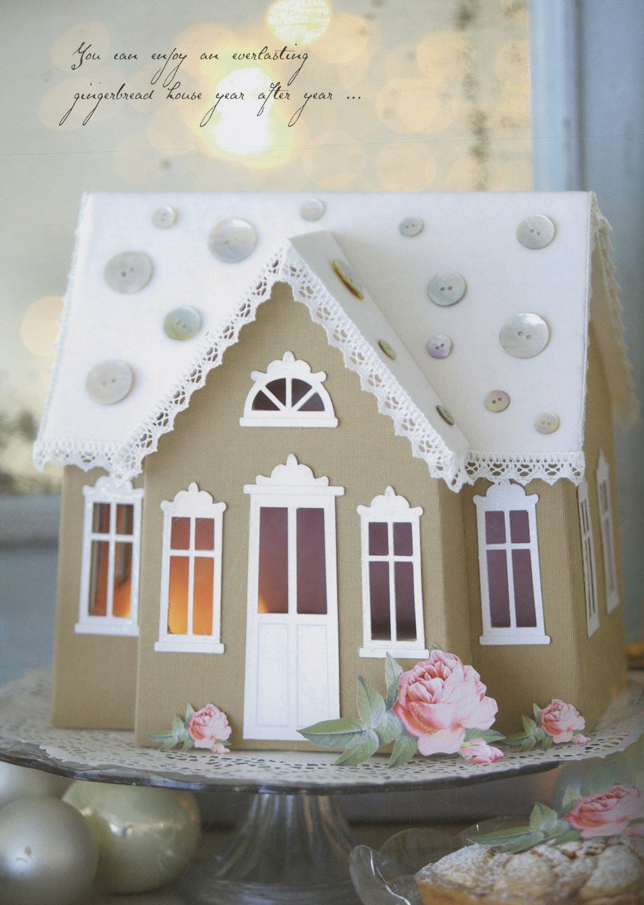 A decorative gingerbread house-shaped cake or candle displayed on a glass pedestal plate. The house has a beige body, white roof and trim, button details on the roof, and pink roses near the base.
Photo by Tildafabrics.com [additional_image_link]