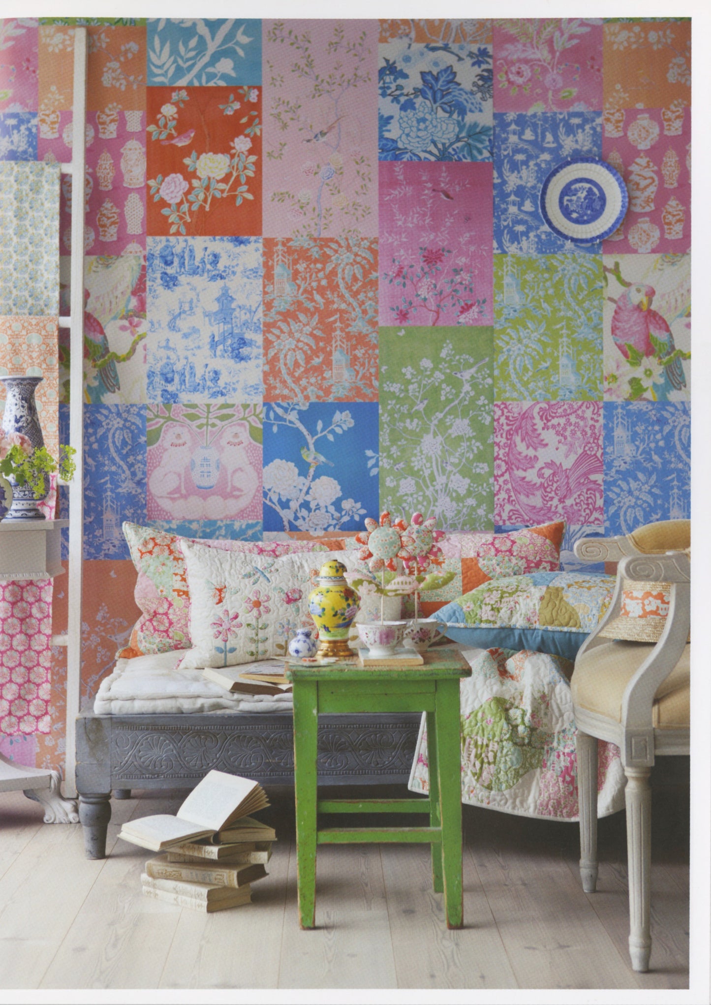 A colorful patchwork wallpaper featuring a grid of floral and botanical patterns in pink, blue, orange, green, and white, styled in a cozy living room setting with a bench, green side table, and patterned pillows.
Photo by Tildafabrics.com [additional_image_link]