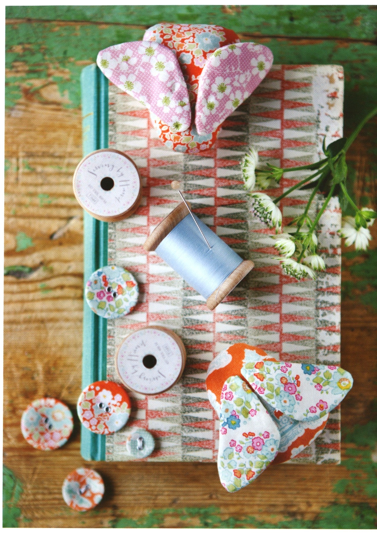 A styled flat-lay of handmade fabric sewing notions, including two pairs of bunny-shaped fabric ears, several floral patterned buttons, a spool of light blue thread, and a decorative book on a rustic wooden surface.
Photo by Tildafabrics.com [additional_image_link]