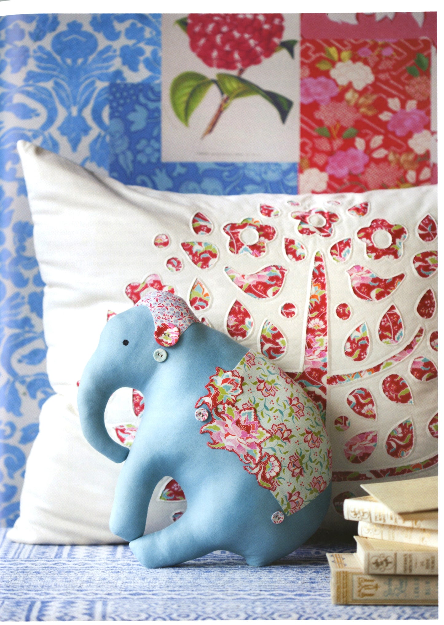 A light blue fabric elephant figurine wearing a pink floral hat and a green floral saddle, posed in front of two decorative pillows with red floral cutout patterns, set against a patchwork wallpaper background.
Photo by Tildafabrics.com [additional_image_link]