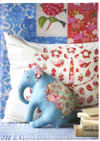 A light blue fabric elephant figurine wearing a pink floral hat and a green floral saddle, posed in front of two decorative pillows with red floral cutout patterns, set against a patchwork wallpaper background.
Photo by Tildafabrics.com [additional_image_link]
