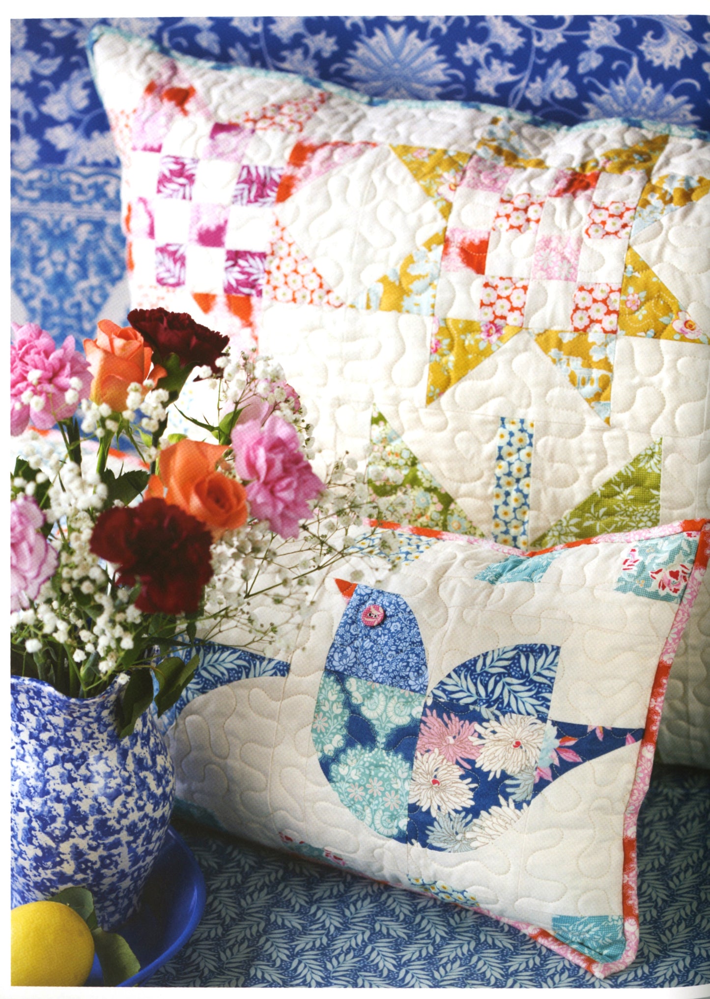 Two quilted patchwork pillows displayed on a blue patterned surface, with a blue-and-white vase of flowers in the foreground. The larger pillow has a multicolor floral patchwork design, and the smaller pillow features a blue bird applique on a white quilted background.
Photo by Tildafabrics.com [additional_image_link]