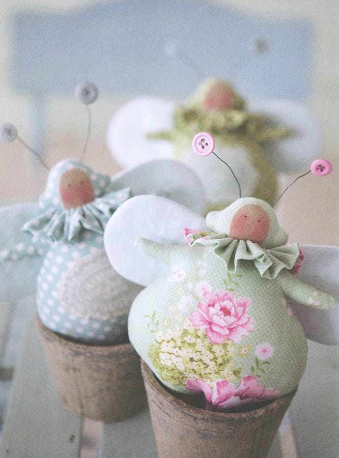 Three decorative fabric pincushion angels with white wings, floral and polka dot patterns, and button antennae, displayed in rustic brown cups.
Photo by Tildafabrics.com [additional_image_link]