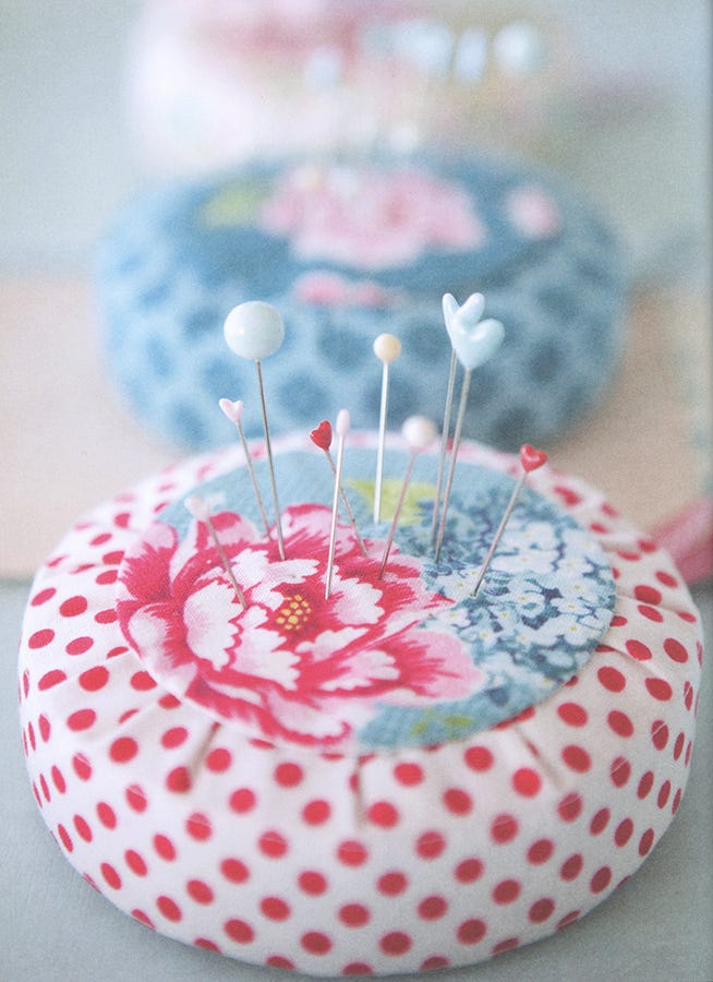 A close-up of a round pin cushion with a red polka-dot and pink floral fabric design, holding several decorative pins, with another floral pin cushion blurred in the background.
Photo by Tildafabrics.com [additional_image_link]