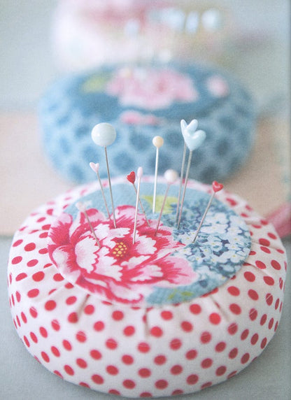 A close-up of a round pin cushion with a red polka-dot and pink floral fabric design, holding several decorative pins, with another floral pin cushion blurred in the background.
Photo by Tildafabrics.com [additional_image_link]