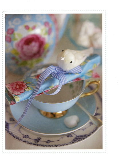 A decorative teacup and saucer set with a light blue porcelain cup and matching saucer, gold trim, floral napkins, a blue-and-white ribbon, and a small white bird figurine on a tabletop.
Photo by Tildafabrics.com [additional_image_link]