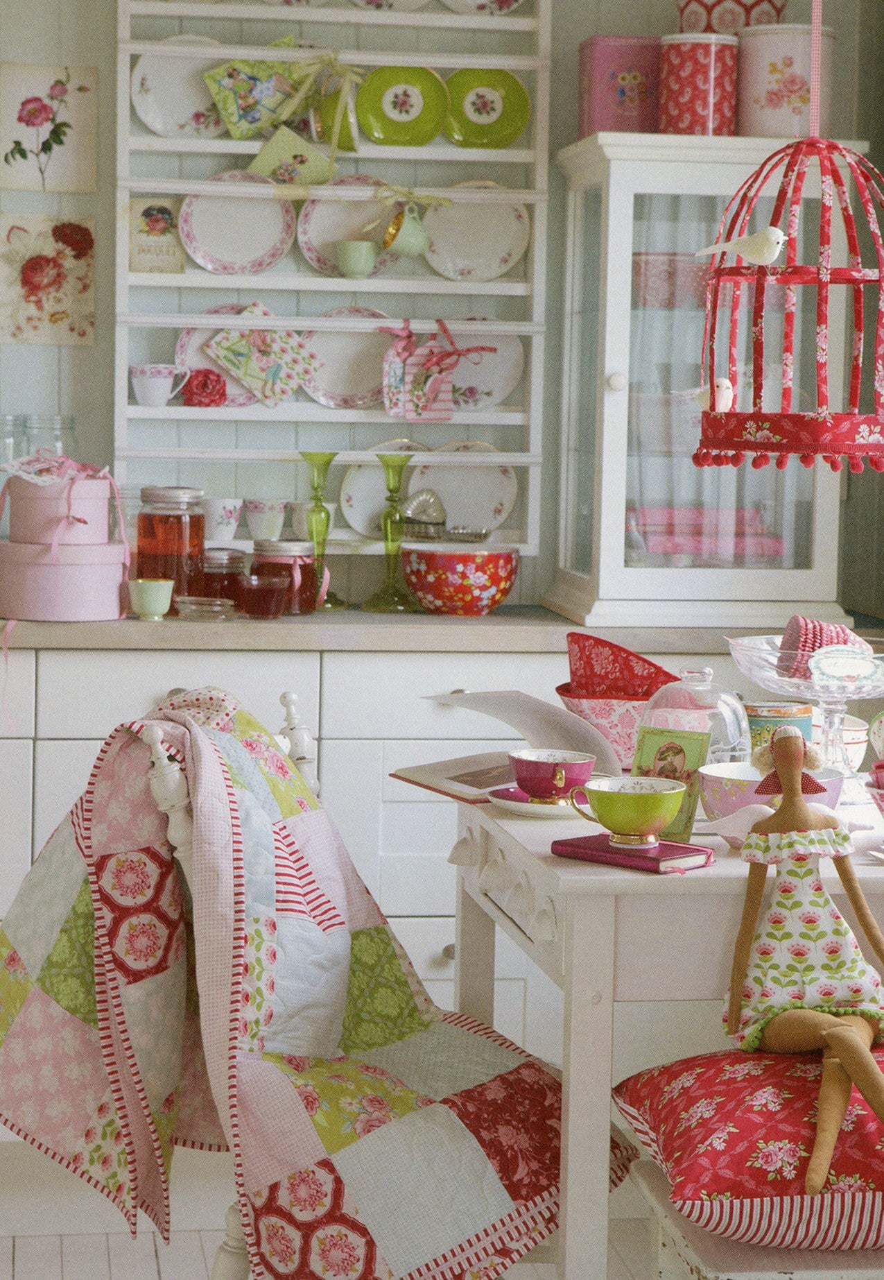 A cozy, vintage-style kitchen scene featuring a white wooden table and chairs decorated with red and green floral quilts, a doll, and teacups, set against a background of white cabinets and shelves filled with dishes and jars.
Photo by Tildafabrics.com [additional_image_link]