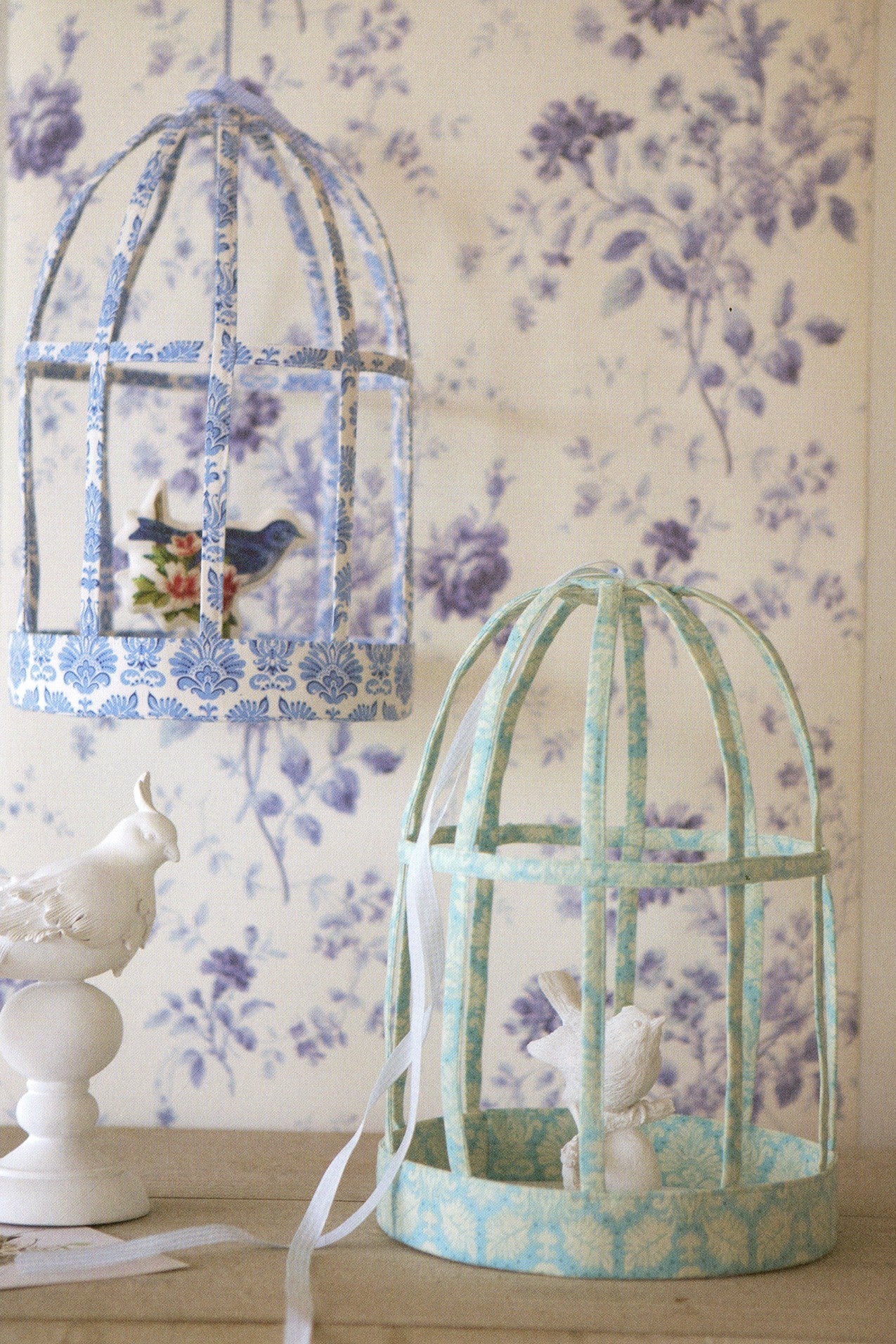 Two decorative birdcage-style hanging domes with blue and mint floral patterned trim, displayed against a purple floral wallpaper background. One dome is open with a small bird figurine inside, and the other is closed with a white bunny figurine.
Photo by Tildafabrics.com [additional_image_link]