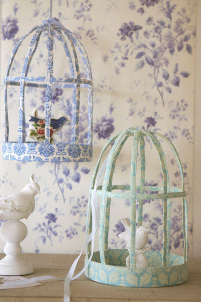 Two decorative birdcage-style hanging domes with blue and mint floral patterned trim, displayed against a purple floral wallpaper background. One dome is open with a small bird figurine inside, and the other is closed with a white bunny figurine.
Photo by Tildafabrics.com [additional_image_link]