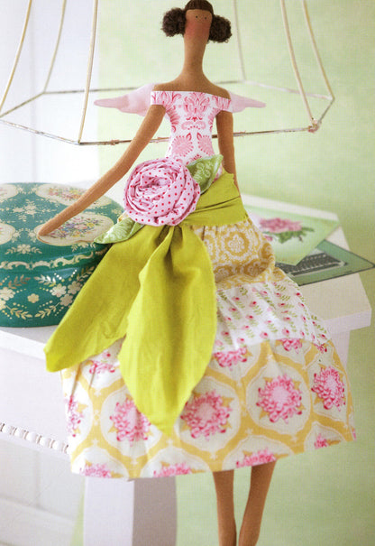 A handmade fabric doll with brown hair and a patterned dress featuring pink, yellow, and white floral prints, posed on a white table against a pale green background.
Photo by Tildafabrics.com [additional_image_link]
