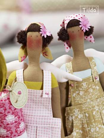 Two handmade fabric dolls with brown paper-like bodies, dark curly hair, pink flower hair accessories, and white wings, wearing patterned aprons. One doll wears a pink-and-white gingham apron over a yellow top, and the other wears a beige-and-white floral apron over a light blue top.
Photo by Tildafabrics.com [additional_image_link]
