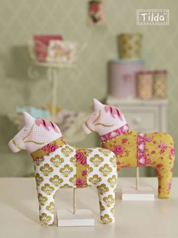 Two decorative fabric-covered wooden doll horses displayed on small stands in a softly styled indoor setting. One horse is white with gold floral motifs and pink accents, and the other is yellow with pink floral print.
Photo by Tildafabrics.com [additional_image_link]