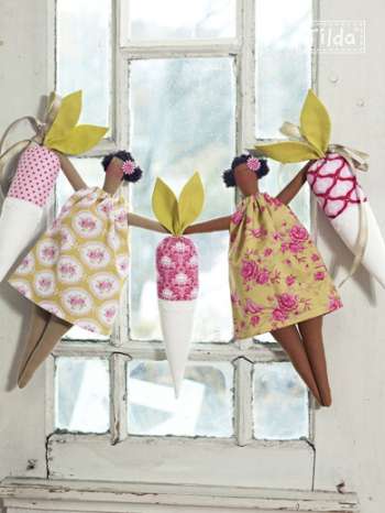 Three handmade fabric carrot dolls with patterned dresses hang in front of a white window. Each doll has a different floral and geometric dress design in pink, yellow, and white, with yellow fabric leaves and brown wooden legs.
Photo by Tildafabrics.com [additional_image_link]