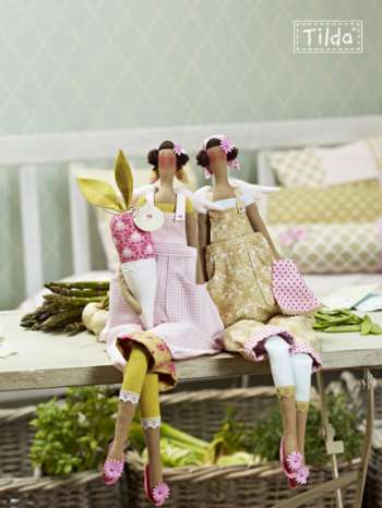 Two handmade fabric dolls dressed in vintage-style gardening outfits, sitting on a rustic wooden table with asparagus and greenery in the background. The brand name 'Tilda' is visible in the top right corner.
Photo by Tildafabrics.com [additional_image_link]