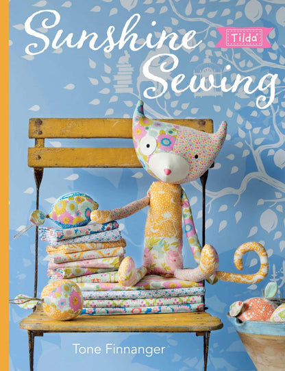 Front cover of the sewing book 'Sunshine Sewing' by Tone Finnanger, featuring a colorful handmade stuffed cat doll sitting on a wooden chair with folded fabric pieces around it against a blue patterned background.
Photo by Tildafabrics.com [image_link]