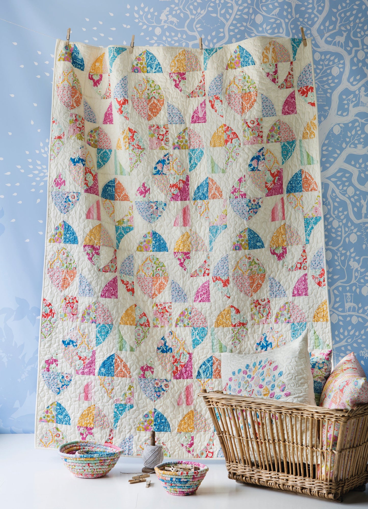 A colorful patchwork quilt hanging vertically in a wicker basket, with matching patterned bowls and a pillow on a white surface against a blue patterned backdrop.
Photo by Tildafabrics.com [additional_image_link]