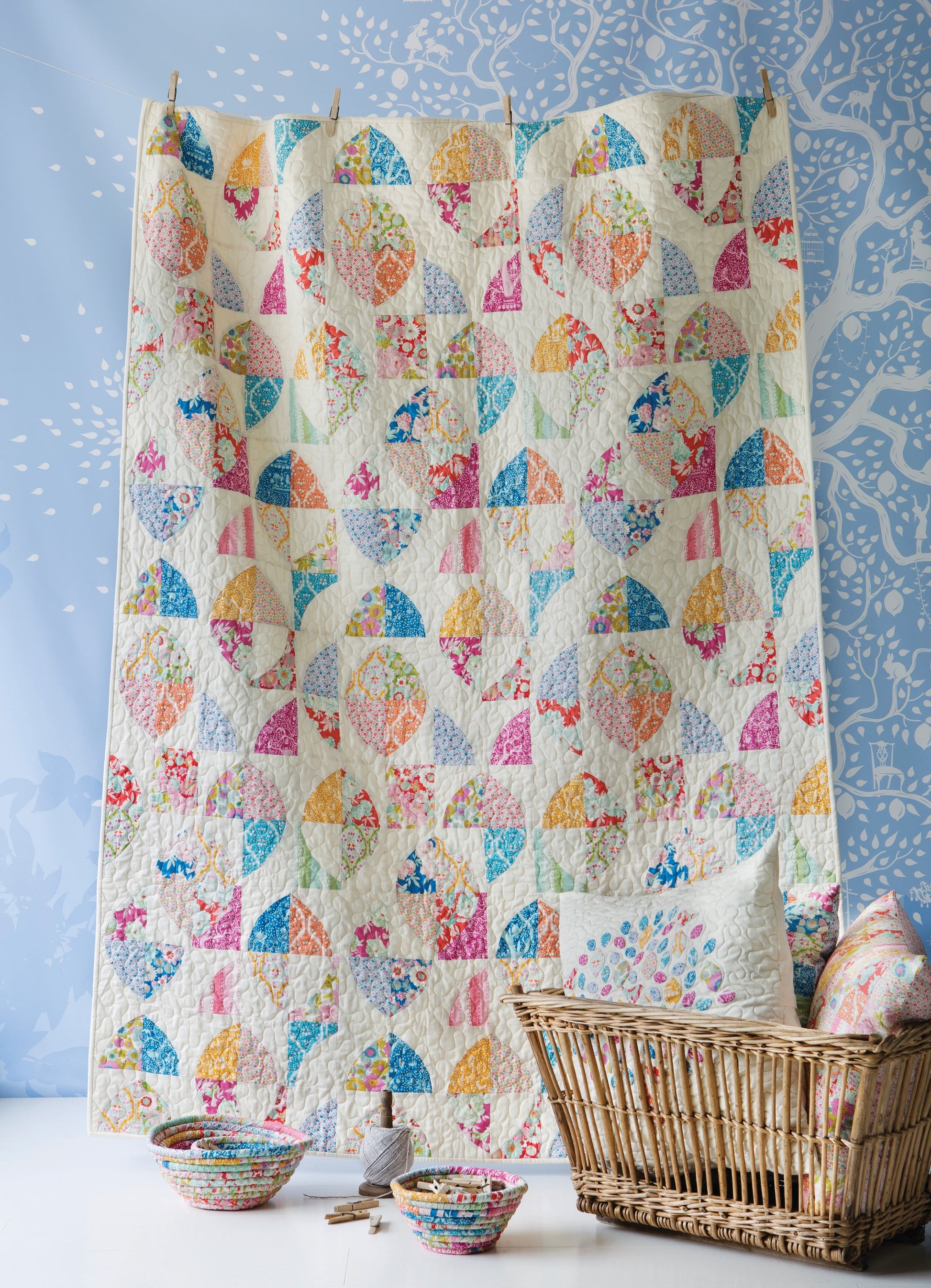A colorful patchwork quilt hanging vertically in a wicker basket, with matching patterned bowls and a pillow on a white surface against a blue patterned backdrop.
Photo by Tildafabrics.com [additional_image_link]