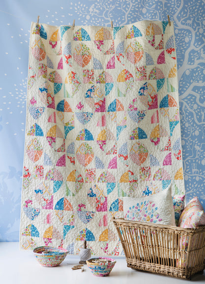 A colorful patchwork quilt hanging vertically in a wicker basket, with matching patterned bowls and a pillow on a white surface against a blue patterned backdrop.
Photo by Tildafabrics.com [additional_image_link]