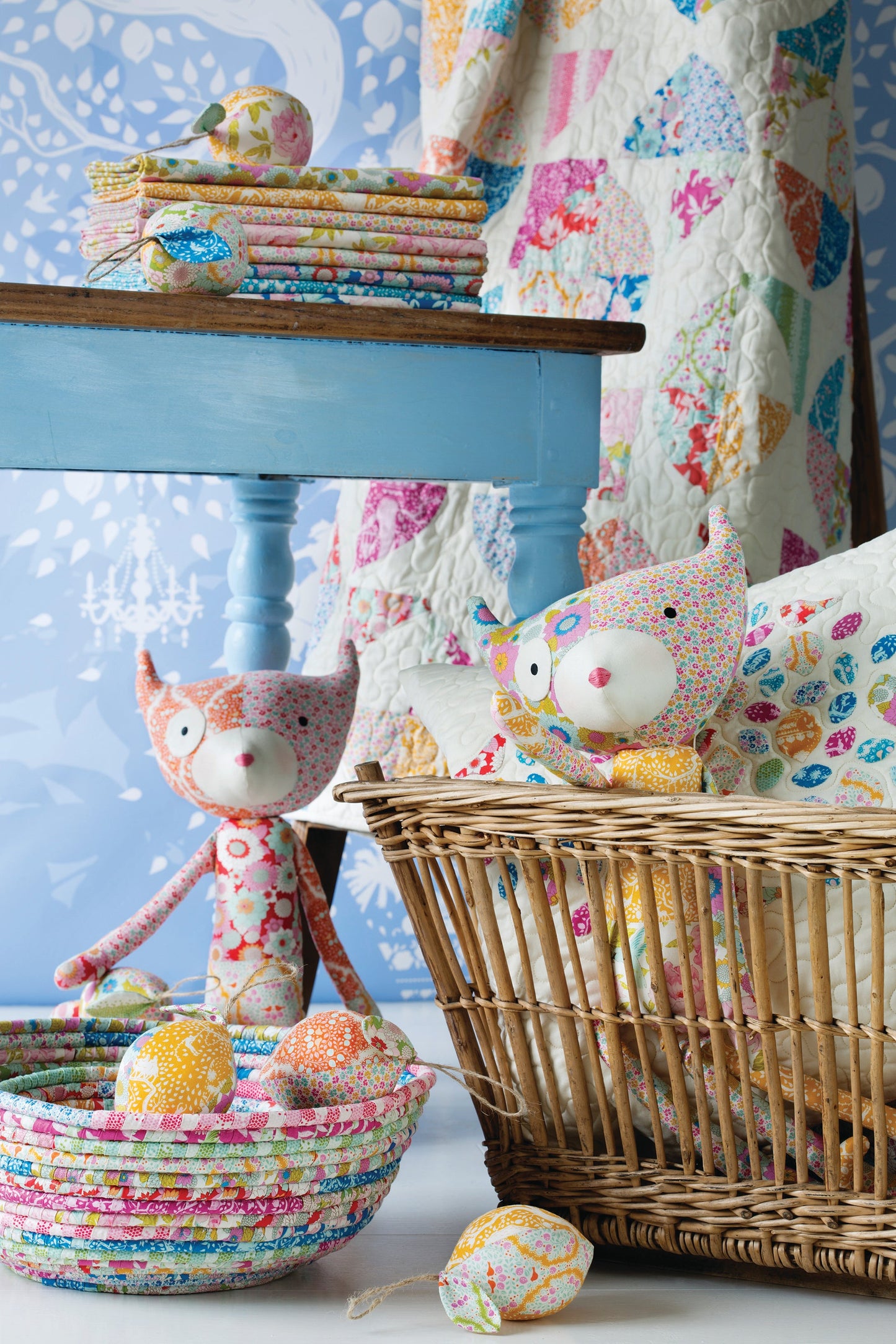 A styled craft display featuring handmade quilted fabric toys, including a pink patterned stuffed owl doll, a striped woven basket, and matching fabric eggs, set against a blue wooden table and a colorful quilted backdrop.
Photo by Tildafabrics.com [additional_image_link]