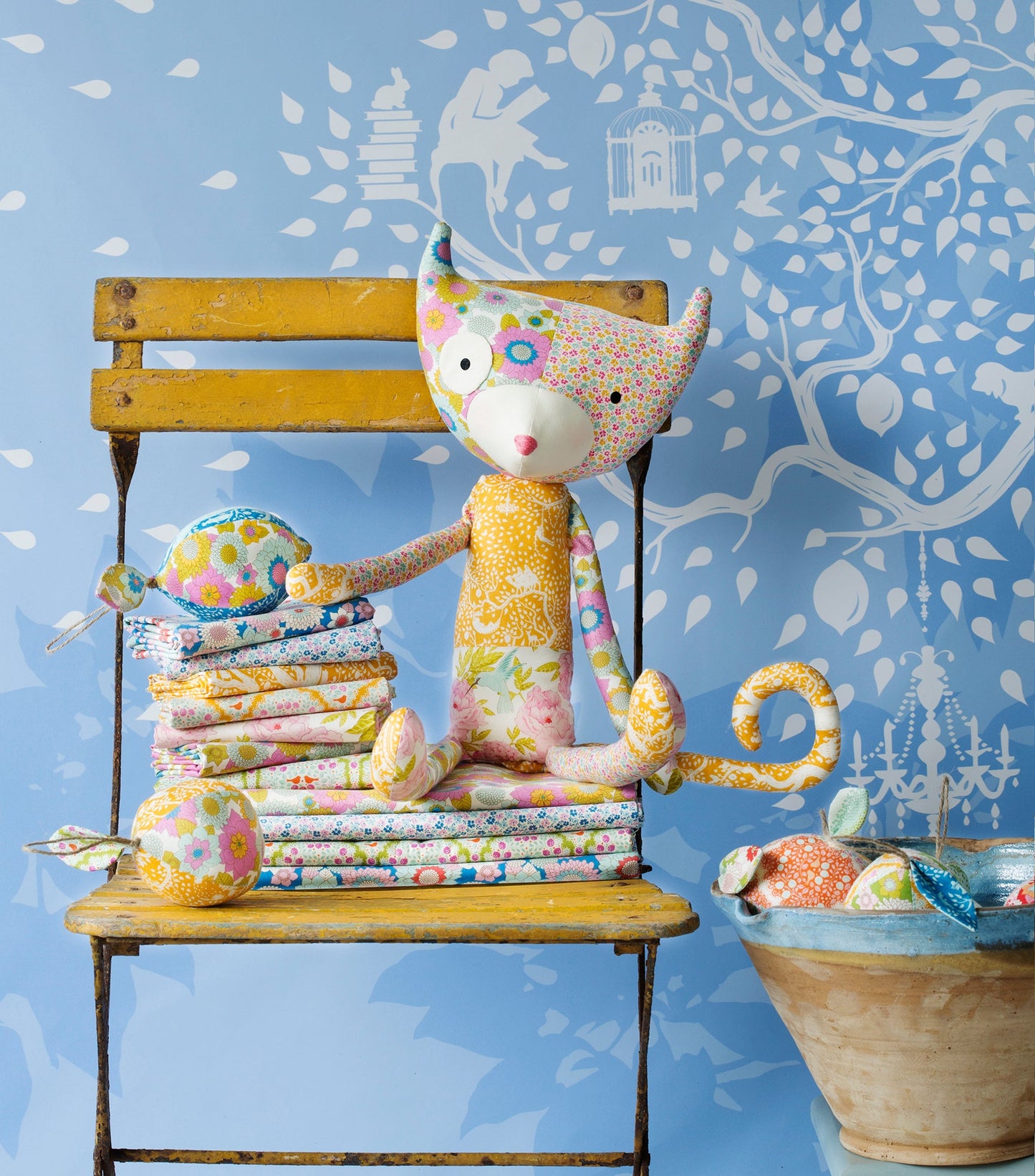 A handmade patchwork fabric cat doll sits on a yellow wooden chair, surrounded by matching patterned fabric rolls and a bowl, set against a blue patterned wallpaper background.
Photo by Tildafabrics.com [additional_image_link]