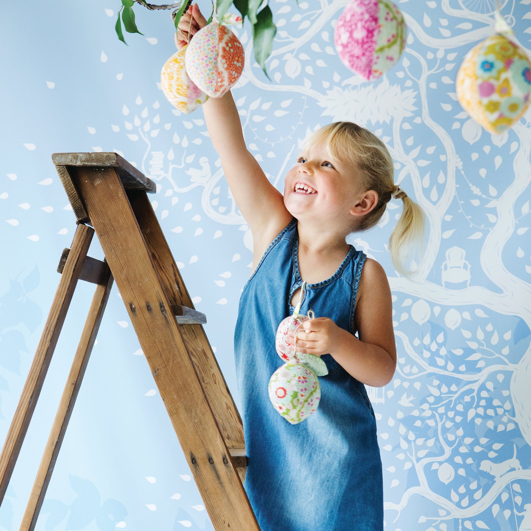 A young blonde girl in a blue denim dress stands on a wooden ladder, reaching for colorful patterned Easter eggs hanging from a tree branch against a light blue wall with white tree illustrations. She holds a matching patterned egg in her hand.
Photo by Tildafabrics.com [additional_image_link]