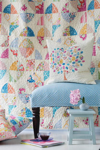 A colorful quilted wall hanging or tapestry with patchwork fan and leaf motifs in pink, blue, yellow, orange, and green, styled in a room scene behind a blue-and-white polka dot bench with a matching decorative pillow.
Photo by Tildafabrics.com [additional_image_link]