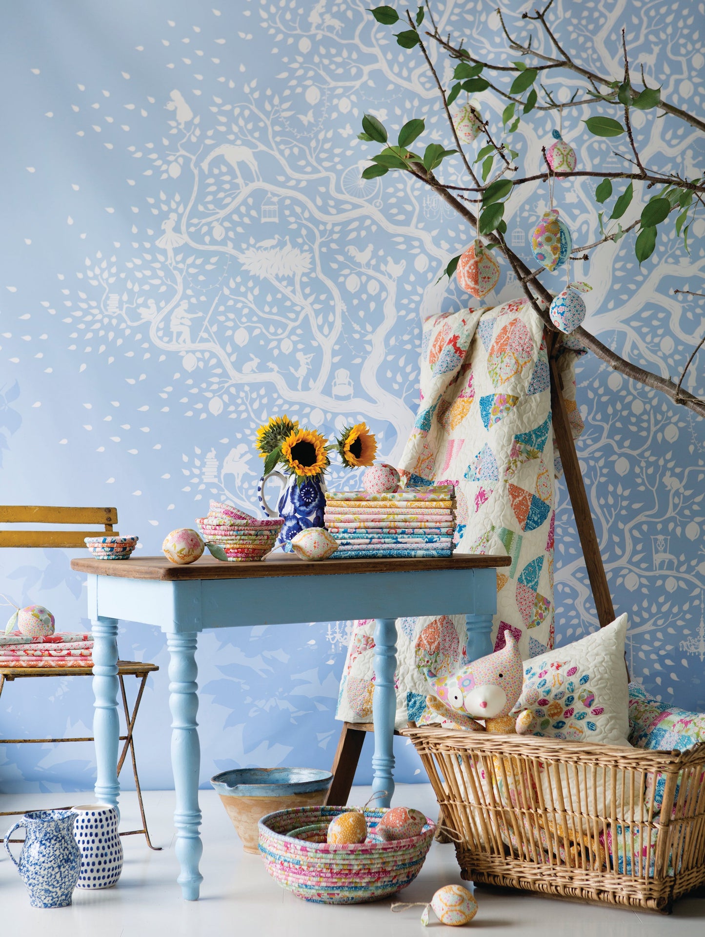 A styled interior scene with a light blue table, pastel quilt draped over a wooden stand, patterned cushions in a wicker basket, sunflowers, eggs, and a blue patterned wallpaper background.
Photo by Tildafabrics.com [additional_image_link]