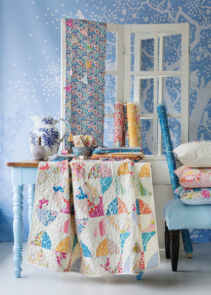 A styled interior scene showing a quilted patchwork textile draped over a table, with matching folded quilts, fabric rolls, and a folded textile panel leaning against a white window frame. The room has blue patterned wallpaper, a blue chair, and decorative ceramic and fabric accessories.
Photo by Tildafabrics.com [additional_image_link]