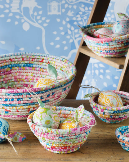 A collection of handmade fabric basketweave bowls in various sizes, decorated with colorful patterned fabric and filled with Easter eggs, arranged on a wooden table and a wooden ladder against a blue patterned wallpaper background.
Photo by Tildafabrics.com [additional_image_link]