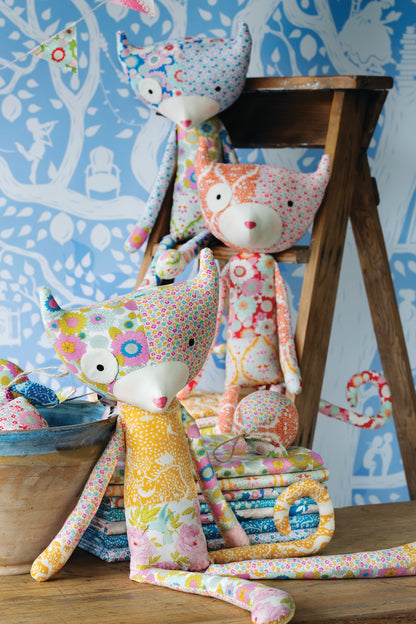 Three handmade fabric fox dolls in colorful floral and polka-dot patterns are arranged on a wooden stool and tabletop against a blue patterned wallpaper background.
Photo by Tildafabrics.com [additional_image_link]