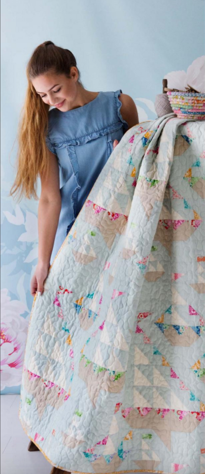 A woman in a blue sleeveless top holds up a large quilted blanket with a light blue base and colorful floral and geometric patterns.
Photo by Tildafabrics.com [additional_image_link]
