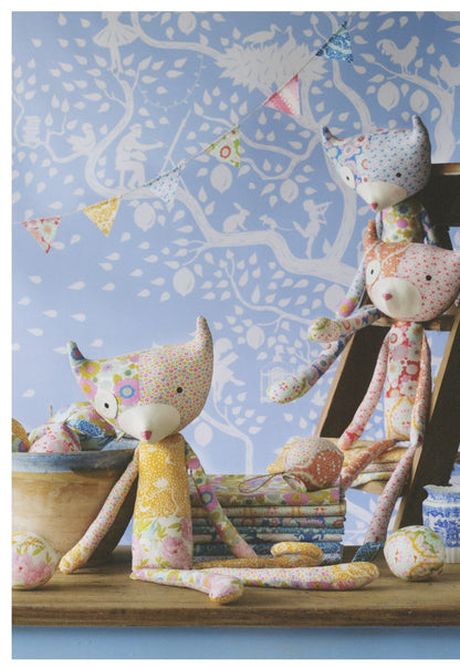 Three handmade fabric fox dolls in colorful floral and polka-dot patterns are arranged on a wooden stool and tabletop against a blue patterned wallpaper background.
Photo by Tildafabrics.com [additional_image_link]