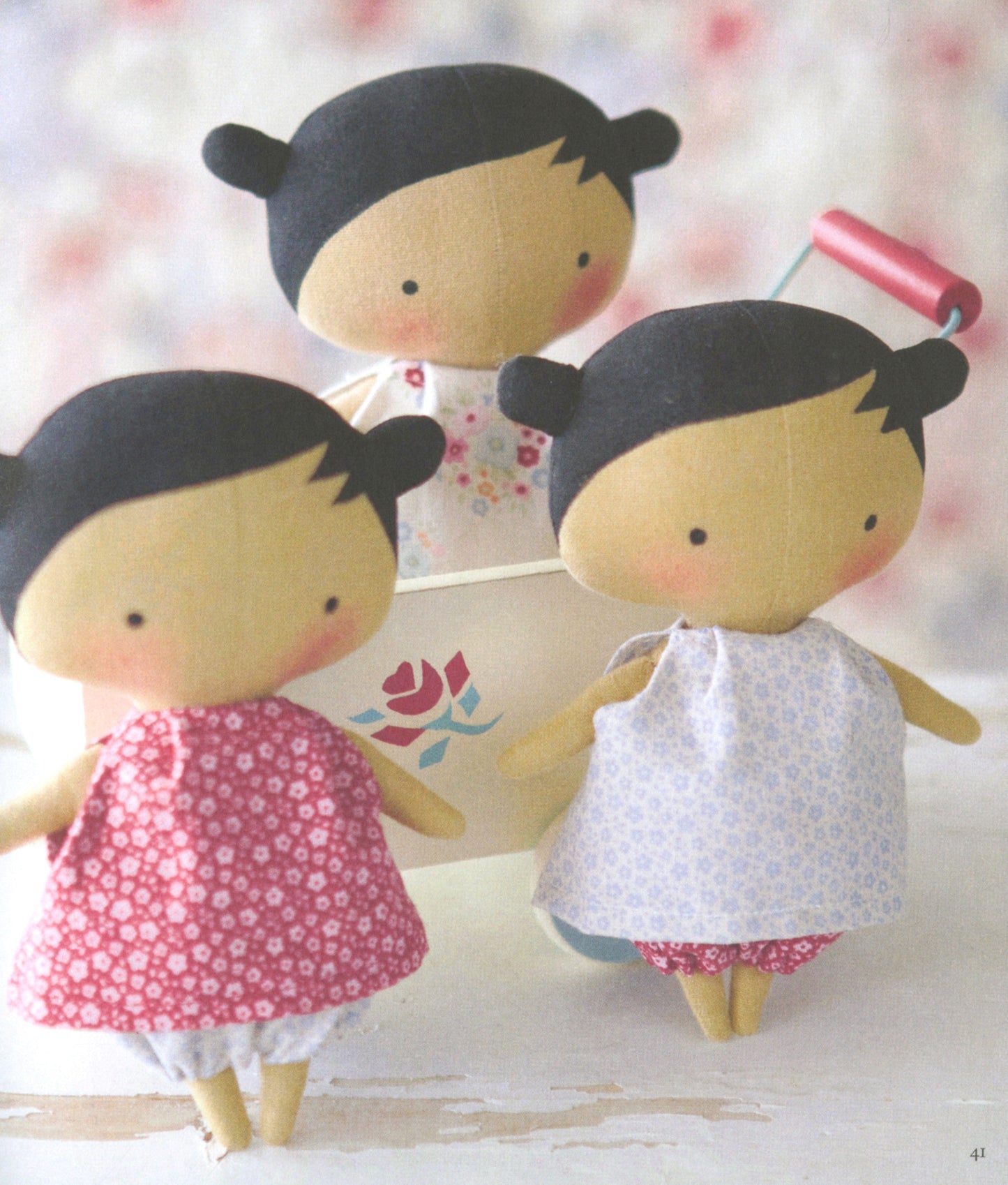 Three handmade fabric doll figures with black hair and pale skin tones, dressed in pink and white floral clothing, posed around a small white flower pot on a soft blurred background.
Photo by Tildafabrics.com [additional_image_link]