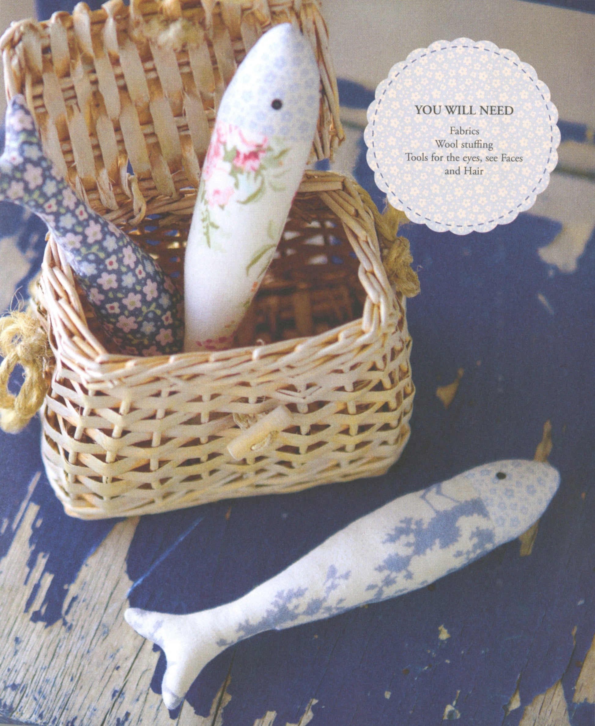 A wicker basket filled with handmade fish-shaped fabric dolls, with one white fish doll featuring pink floral details and another blue-and-white patterned doll partially visible. A circular lace-style text overlay reads 'YOU WILL NEED' with fabric and tools listed.
Photo by Tildafabrics.com [additional_image_link]