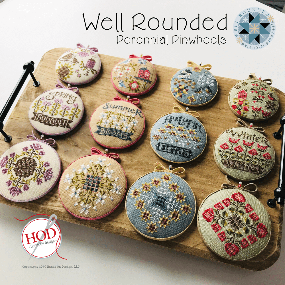 Twelve round embroidered cross-stitch pinwheel ornaments displayed on a wooden board, each with a different seasonal or decorative design and small ribbon loops at the top. The image includes the text 'Well Rounded Perennial Pinwheels' and a 'HOD Hands on Design' logo.
Photo by HandsOnDesign.biz [image_link]