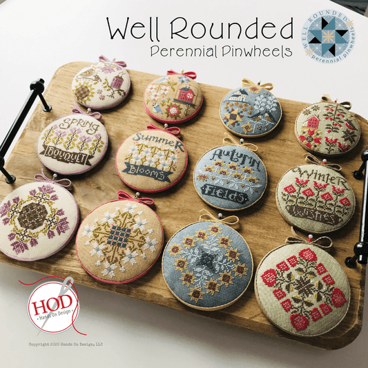 Twelve round embroidered cross-stitch pinwheel ornaments displayed on a wooden board, each with a different seasonal or decorative design and small ribbon loops at the top. The image includes the text 'Well Rounded Perennial Pinwheels' and a 'HOD Hand on Deck Designs' logo.
Photo by HandsOnDesign.biz [image_link]